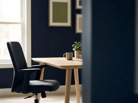 Empty office chair pulled back from a clean wooden desk in a small-business workspace with deep navy walls, muted green accents, and soft natural window light, leaving generous negative space on the right.
