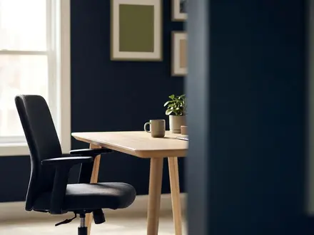 Empty office chair pulled back from a clean wooden desk in a small-business workspace with deep navy walls, muted green accents, and soft natural window light, leaving generous negative space on the right.