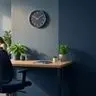 Empty workstation with an unoccupied chair and bare desk in the foreground of a small business office, with blurred coworkers at other desks in the background, in deep navy and green tones to convey the cost of a long-vacant role.